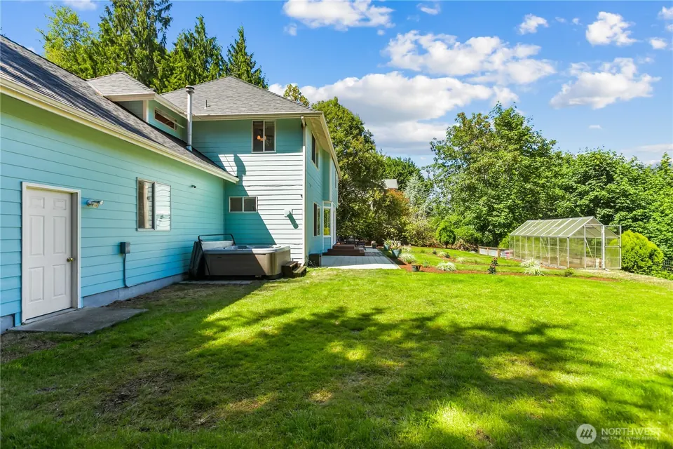 Back of the house with a deck, hot tub and greenhouse on a wide lawn.